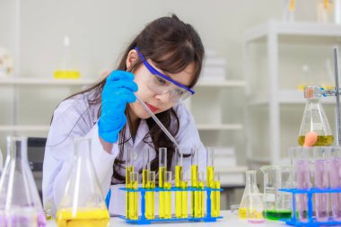 Asyalı genç kadın laboratuvar asistanı profesyonel bilim adamı beyaz laboratuvar önlüğü ve koruyucu gözlüklerle kimya laboratuarında deney tüpleri üzerinde araştırma yapıyor.