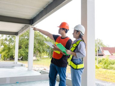 Asyalı erkek ve kadın inşaat işçileri güvenlik donanımı işçisi planlarını tartışıyor, inşaat sahasında takım çalışması planlıyor, pozitiflik, işbirliği başarılı bir proje yürütülüyor..