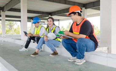 Asian men and women construction workers in safety gear and helmet sitting taking a well deserved break on a bustling job site with blueprints discuss plan working teamwork successful project progress clipart