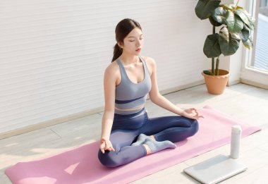 Asyalı genç ve çekici bir kadın spor kıyafetleri giyer yoga minderi çalışır düşüncelere dalarken internetten bilgisayar eğitimi alırken oturma odasında sağlıklı yaşam tarzında..