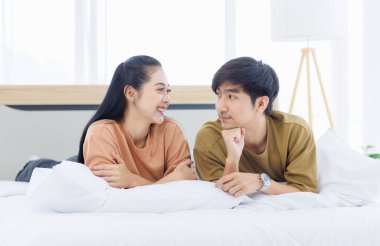 Genç Asyalı sevimli, sevimli, çekici, neşeli, mutlu çiftin portresi gülümsemeyi sever ve yatak odasındaki beyaz yatakta dinlenir. Sevgililer Günü 'nde ve ilişki konseptinde birlikte romantik vakit geçiririz..