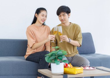 Asyalı genç, mutlu ve çekici çift, sevgililerinin gülümsemesini sever. Ellerinde şampanya bardaklarıyla kadeh tokuştururlar. Oturma odasındaki rahat koltukta oturup yıldönümlerini kutlarlar..