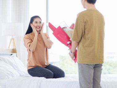 Asyalı, genç, mutlu, çekici bir çift, aşıklar gülüşü bir buket kırmızı gül verir. Beyaz bir yatakta, yatak odasında, sevgililer gününde romantik vakit geçirmenin keyfini çıkarır..
