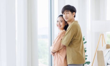 Yan görünümün portresi. Genç, tatlı, romantik Asyalı mutlu çift kucaklaşmayı sever. Gülümseyen kamera, yatak odasındaki perdenin yanında..