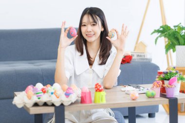 Asyalı, çekici, neşeli, sevimli, hoş, sıradan kıyafetler giyen, neşeli, komik bir gülümseme taşıyan, renkli yumurtalar taşıyan, evde oturmuş, Paskalya kutlaması için hazırlanan bir kadın..