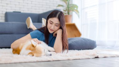 Neşeli Asyalı genç güzel kadın sevimli Galli Corgi köpeğiyle mutlu bir gülümseme ile hayvanlarla poz verirken ev konsepti yaşam tarzının oturma odasında halı üzerinde uzanırken evcil hayvan en iyi arkadaşının keyfini çıkar..