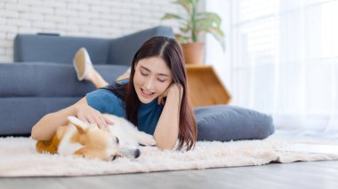 Neşeli Asyalı genç güzel kadın sevimli Galli Corgi köpeğiyle mutlu bir gülümseme ile hayvanlarla poz verirken ev konsepti yaşam tarzının oturma odasında halı üzerinde uzanırken evcil hayvan en iyi arkadaşının keyfini çıkar..