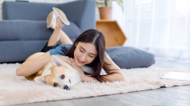 Neşeli Asyalı genç güzel kadın sevimli Galli Corgi köpeğiyle mutlu bir gülümseme ile hayvanlarla poz verirken ev konsepti yaşam tarzının oturma odasında halı üzerinde uzanırken evcil hayvan en iyi arkadaşının keyfini çıkar..