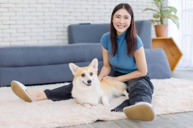 Asyalı genç ve güzel bir kadın mutlu bir gülümseme, şirin bir Galli mantar köpeği ile evde halı döşemesinde komik bir şekilde oturuyor, arkadaşlık hayatı konsepti, sevgi dolu hayvanlarla vakit geçirmenin keyfini çıkarıyor.