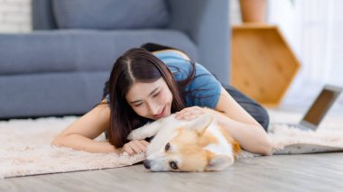 Neşeli Asyalı genç güzel kadın sevimli Galli Corgi köpeğiyle mutlu bir gülümseme ile hayvanlarla poz verirken ev konsepti yaşam tarzının oturma odasında halı üzerinde uzanırken evcil hayvan en iyi arkadaşının keyfini çıkar..