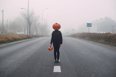 Cadılar Bayramı. Balkabağı. Kafasında balkabağı olan bir adam sisli bir caddenin ortasında duruyor. Elinde bir balkabağı feneri tutuyor.