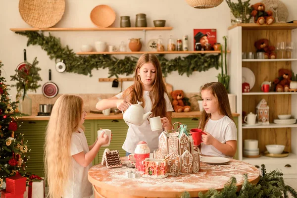 Three pretty young girls, sisters, prepared a Christmas gingerbread house in the kitchen. They are standing at a round table and are going to drink tea. New Year, Christmas.