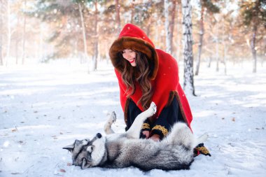 Kış ormanı ve kar yağışının arka planında gri köpekli kırmızı başlıklı genç bir kadın..