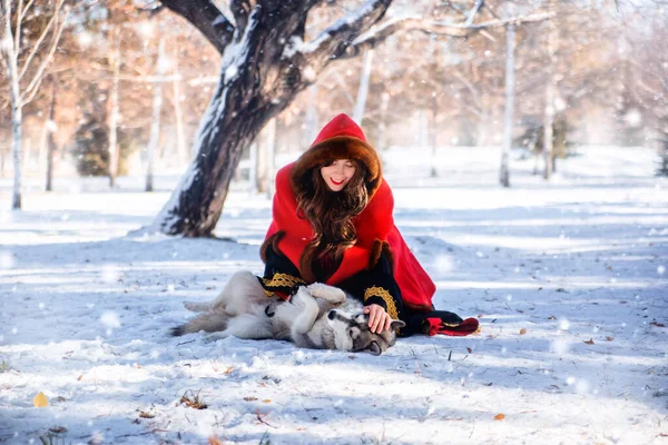 冬の森や雪を背景に灰色のハスキー犬と赤いフード付きのレインコートの若い女性 — ストック写真