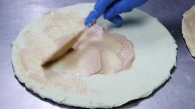 Close-up of the hands of the cook in blue gloves spread sauce, ketchum, mayonnaise on the pizza crust with a kitchen spatula.