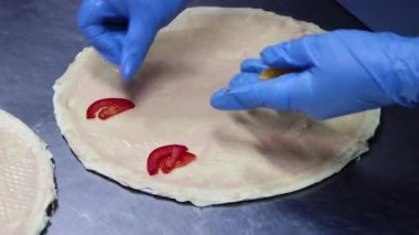 The process of making pizza, smooth camera movement, selective focus, close-up of the chef's hands in blue gloves lay out vegetables, tomatoes on a pizza crust smeared with sauce, ketchum, mayonnaise.