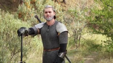 Ortaçağ savaşçısı portresi, gri saçlı, yüzünde zincirli bir yara izi, omuzlarında vatkalarla ormanın zeminine karşı duran iki elli bir kılıç. Orta Çağ Cadısı