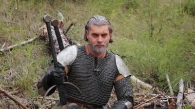 Düz kamera hareketi, seçici odak, gri saçlı ve yüzünde yara izi olan bir adam elinde iki eliyle devrilmiş bir ağaçta oturuyor. Ortaçağ savaşçısı, yarım zincirlenmiş cadı..