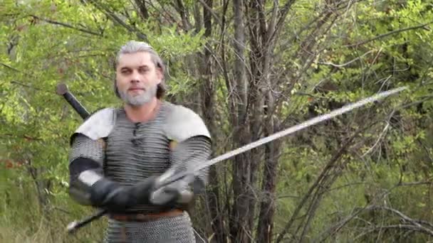 灰色の髪をした中世の戦士と肩パッドと彼の手の剣とチェーンメールで彼の顔に傷は 森の背景に保護 戦闘位置に立っている ウィッチャー — ストック動画