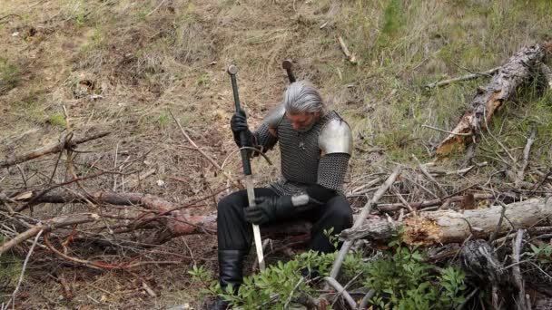 镜头平稳的移动 有选择的焦点 一个头发灰白 脸上有疤的头戴链子的男人坐在倒下的树上擦拭 磨尖了一把双手的剑 中世纪的战士 魔法师 — 图库视频影像
