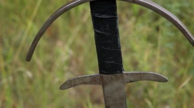 Pürüzsüz kamera hareketi, seçici odaklanma, yerde sıkışmış iki elli bir kılıç bir tarlanın, ormanın ve gökyüzünün arka planında. Kayıp, unutulmuş kılıç, düşmüş savaşçı.