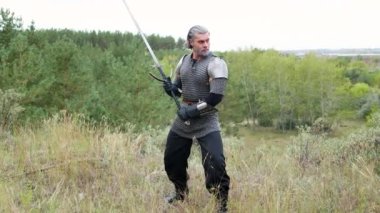 Ortaçağ savaşçısı, gri saçlı ve yüzünde zincirli bir yara izi olan omuzlarında vatkaları ve ellerinde bir kılıçla ormanın zeminine karşı koruyucu ve savaş pozisyonunda duruyor. Cadı