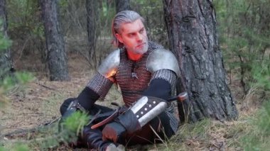 Bir ortaçağ savaşçısının portresi, gri saçlı, yüzünde zincirli bir yara izi omuzluk ve elinde iki elli bir kılıç ile bir ağacın yanında oturuyor. Orta Çağ Cadısı