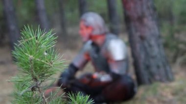 A sharp change of focus from a spruce branch to a portrait of a medieval warrior with gray hair and a scar on his face, dressed in chain mail and holding a two-handed sword, sitting in the forest.