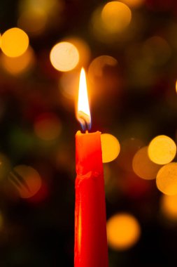 Close-up of a red burning candle in the dark against the bokeh background of a Christmas tree.