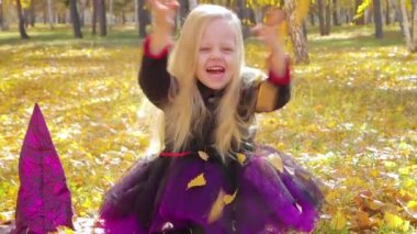 A little girl in a witch costume sits on the grass in the park and catches falling yellow leaves with her hands, smiling broadly.