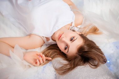 Portrait of an attractive young woman in a white tunic with large white wings behind her back, posing lying on white glowing clouds.