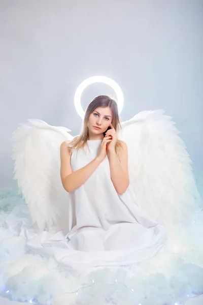 An attractive young woman in a white tunic with large white wings behind her back and a halo above her head poses while sitting in white luminous clouds.