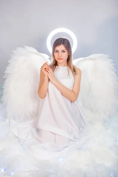 An attractive young woman in a white tunic with large white wings behind her back and a halo above her head poses while sitting in white luminous clouds.