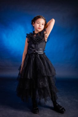 A little girl in a black dress with a pigtail hairstyle on her head poses, isolated on a dark background with blue backlight.