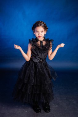 A little girl in a black dress with a pigtail hairstyle on her head poses, isolated on a dark background with blue backlight.