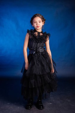 A little girl in a black dress with a pigtail hairstyle on her head poses, isolated on a dark background with blue backlight.