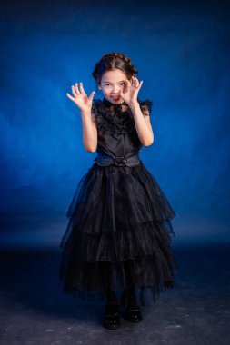 A little girl in a black dress with a pigtail hairstyle on her head poses, isolated on a dark background with blue backlight.