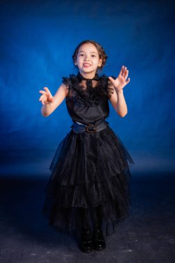 A little girl in a black dress with a pigtail hairstyle on her head poses, isolated on a dark background with blue backlight.
