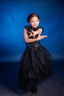 A little girl in a black dress with a pigtail hairstyle on her head poses, isolated on a dark background with blue backlight.