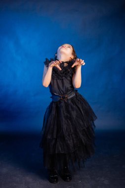 A little girl in a black dress with a pigtail hairstyle on her head poses, isolated on a dark background with blue backlight.