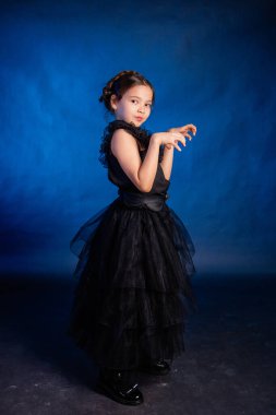 A little girl in a black dress with a pigtail hairstyle on her head poses, isolated on a dark background with blue backlight.