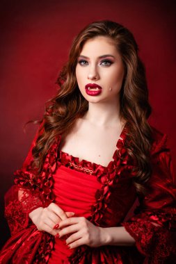 Portrait of a young, attractive vampire woman in a red rococo dress posing isolated against a dark background with red backlights.