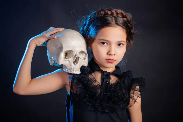 彼女の頭の上にピグテールの髪型と黒のドレスの女の子は 彼女の手の中に頭蓋骨でポーズ青のバックライトで暗い背景に隔離 — ストック写真