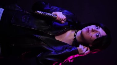 Dikey video, elinde katana olan siyah deri ceketli kısa saçlı bir kadının portresi. Mavi ve mor arka planda izole edilmiş bir şekilde poz veriyor. Siber punk konsepti..