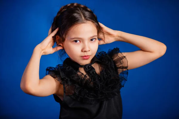 stock image Portrait of a little girl in a black dress with a pigtail hairstyle on her head poses, isolated on a dark background with blue backlight. 