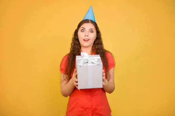stock image Portrait of a young woman in a paper cap with a gift in her hands isolated on a yellow background, birthday concept.