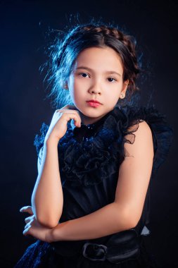 Kafasında örgülü saç stili olan siyah elbiseli küçük bir kızın portresi, koyu bir arka planda, mavi arka planda izole edilmiş.. 