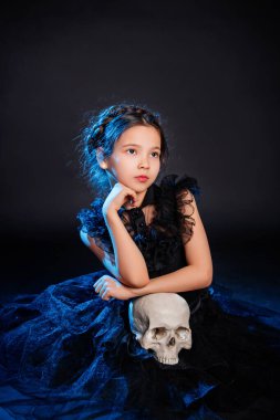 Saçları örgülü siyah elbiseli küçük bir kız ellerinde kafatası, koyu bir arka planda mavi bir ışıkla oturuyor..