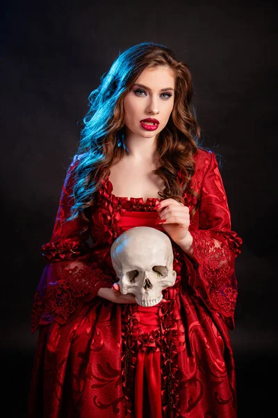 ロココ時代の赤いドレスを着た若い魅力的な吸血鬼の女性の肖像 青いバックライトで暗い背景に隔離された彼女の手の中に頭蓋骨とポーズ — ストック写真