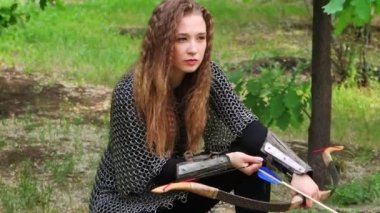 Pürüzsüz kamera hareketi, seçici odaklanma, zincirli zırhlı genç bir kadın ve elinde yayıyla bir yaz ormanı zemininde oturuyor..